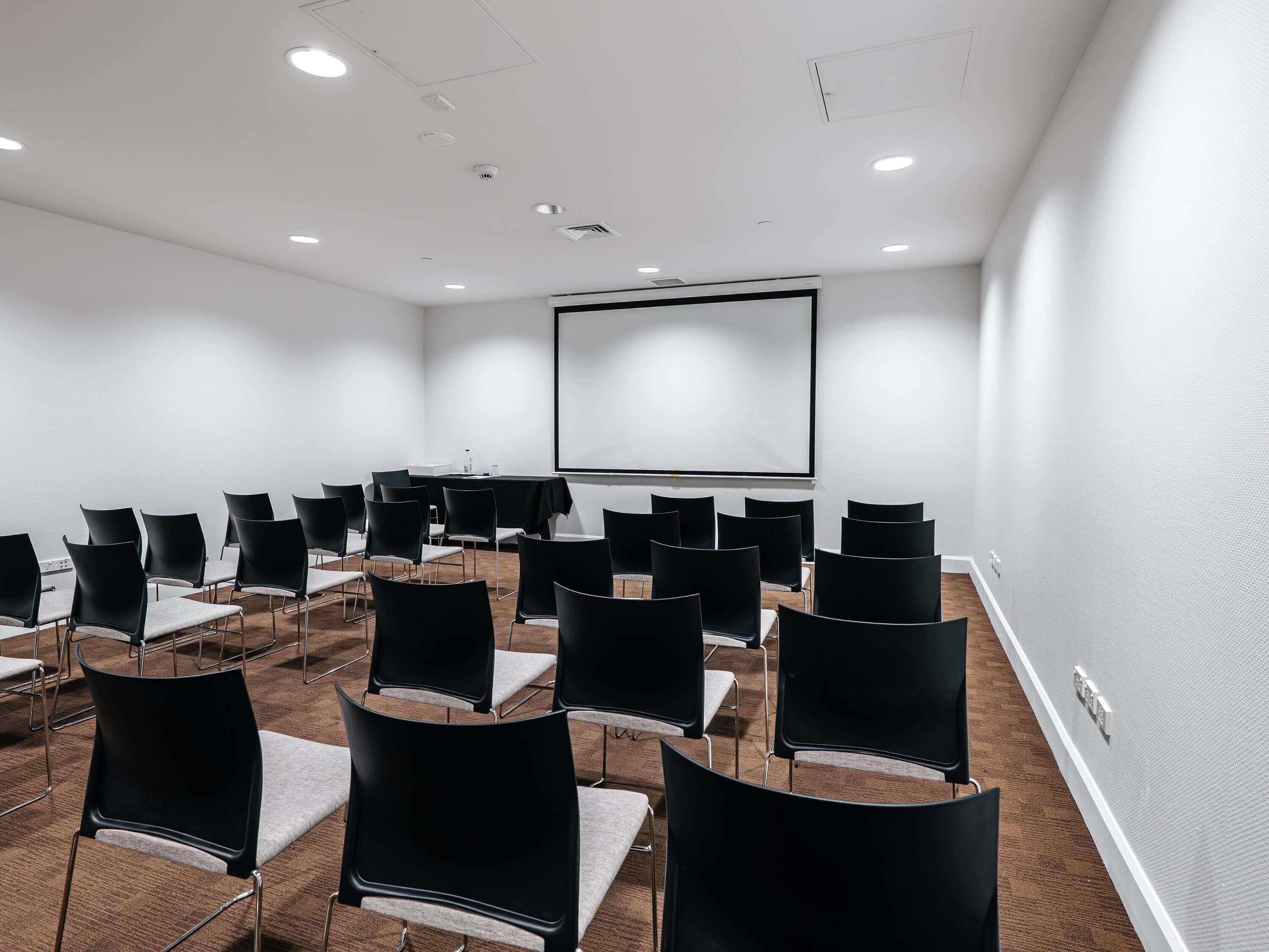 Un salón de conferencias con filas de sillas, una pantalla de proyector y luces en el techo.