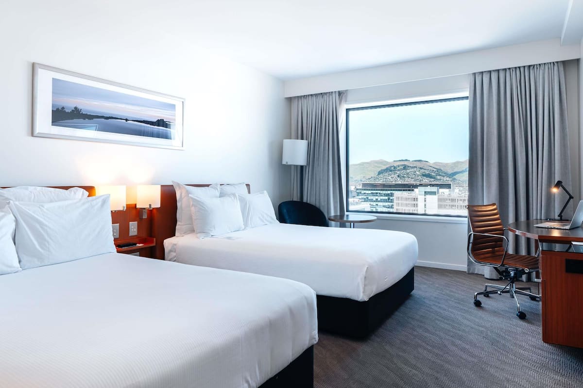 Ein Hotelzimmer mit zwei Betten, einem Schreibtisch mit einem Laptop, einem Stuhl und einem großen Fenster mit Blick auf die Stadt.