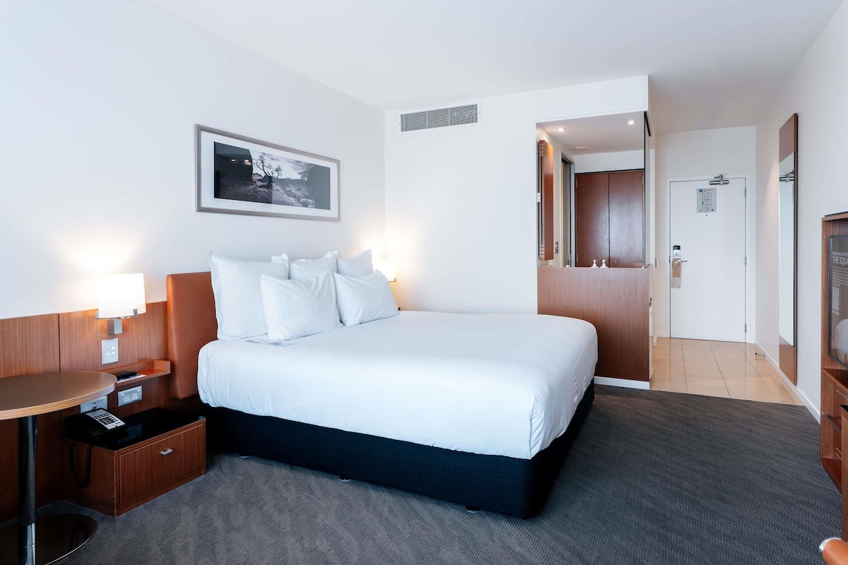 Ein Hotelzimmer mit einem großen Bett, einem Schreibtisch mit Telefon, einem Sessel, einem Wandbild und einer Tür zu einem anderen Raum.