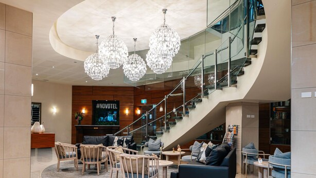 A modern hotel lobby with a curved staircase, chandeliers, seating areas, and a TV screen displaying #NOVOTEL. - Novotel Christchurch Cathedral Square (Christchurch)