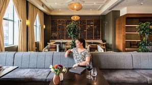 A woman seated on a grey sofa in a well-lit, modern lobby with a large window, a bookshelf, and a decorative ceiling light. - Novotel Christchurch Cathedral Square (Christchurch)