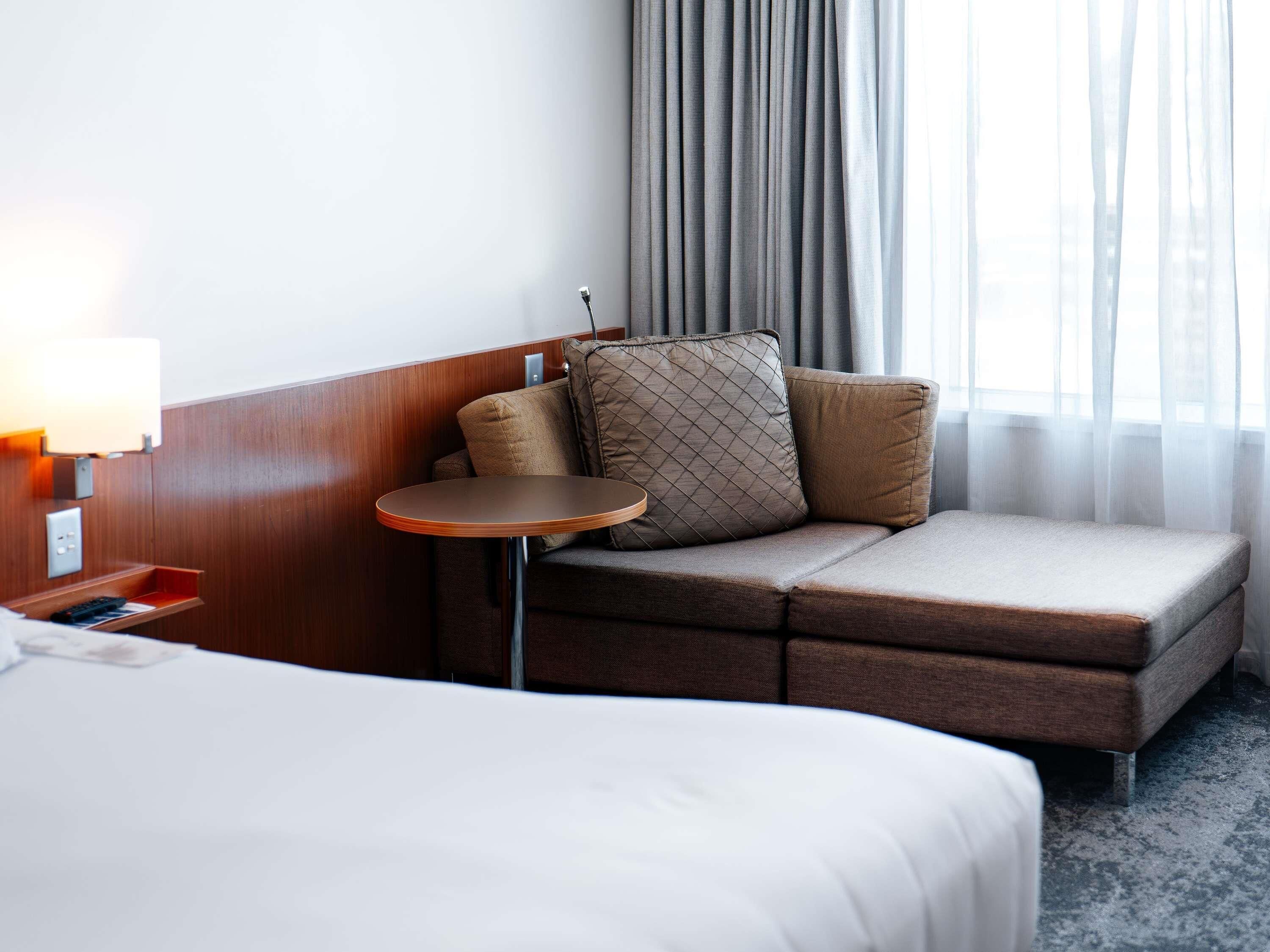 Una habitación de hotel con cama, una mesita redonda pequeña, un sofá con cojines, una lámpara y una ventana con cortinas.