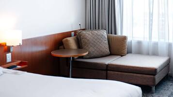 Una habitación de hotel con cama, una mesita redonda pequeña, un sofá con cojines, una lámpara y una ventana con cortinas.