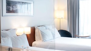 A hotel room with two beds, a bedside table with a lamp, a small round table, a bathroom with a sink and toiletries, and a painting on the wall. - Novotel Christchurch Cathedral Square (Christchurch)