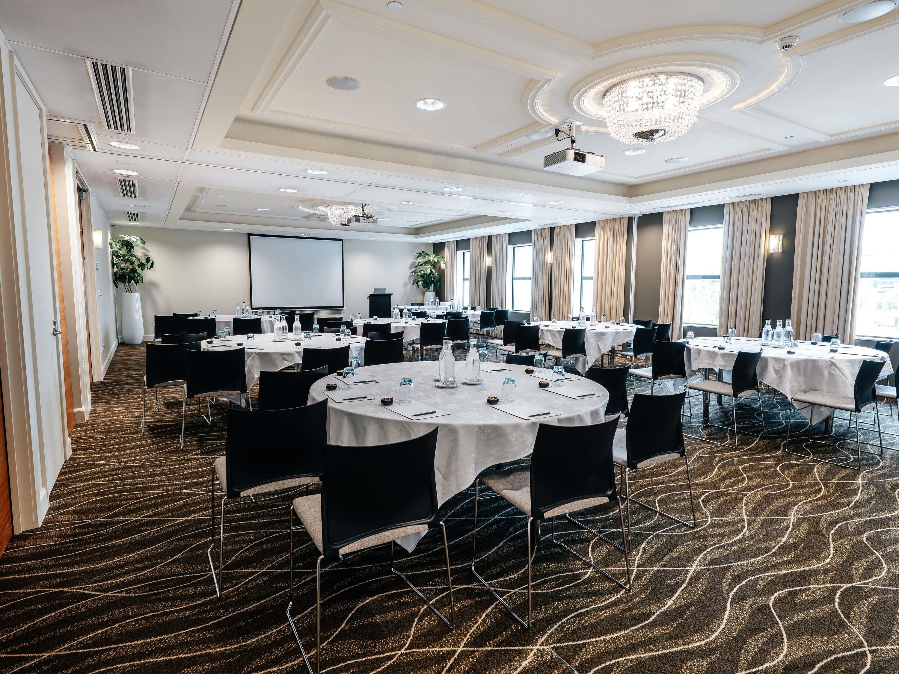 Un salón de conferencias con mesas redondas, sillas, una pantalla de proyector y una planta grande.