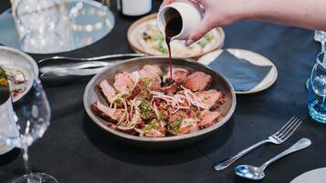 Una mesa de comedor con un plato de carne en rodajas adornado con cebollas y hierbas, una mano sirviendo salsa y varios cubiertos.