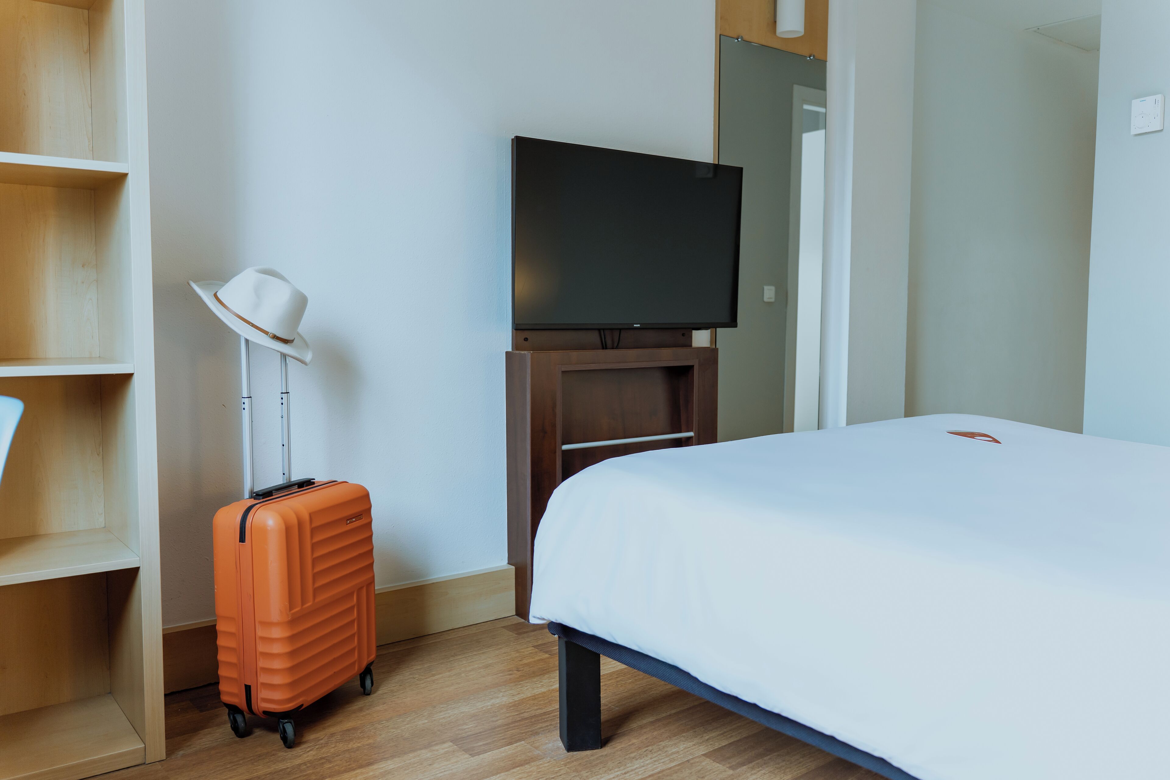 Hotellihuone, jossa on sänky, jalustalla oleva televisio, matkalaukku, hattu ja puinen hylly.