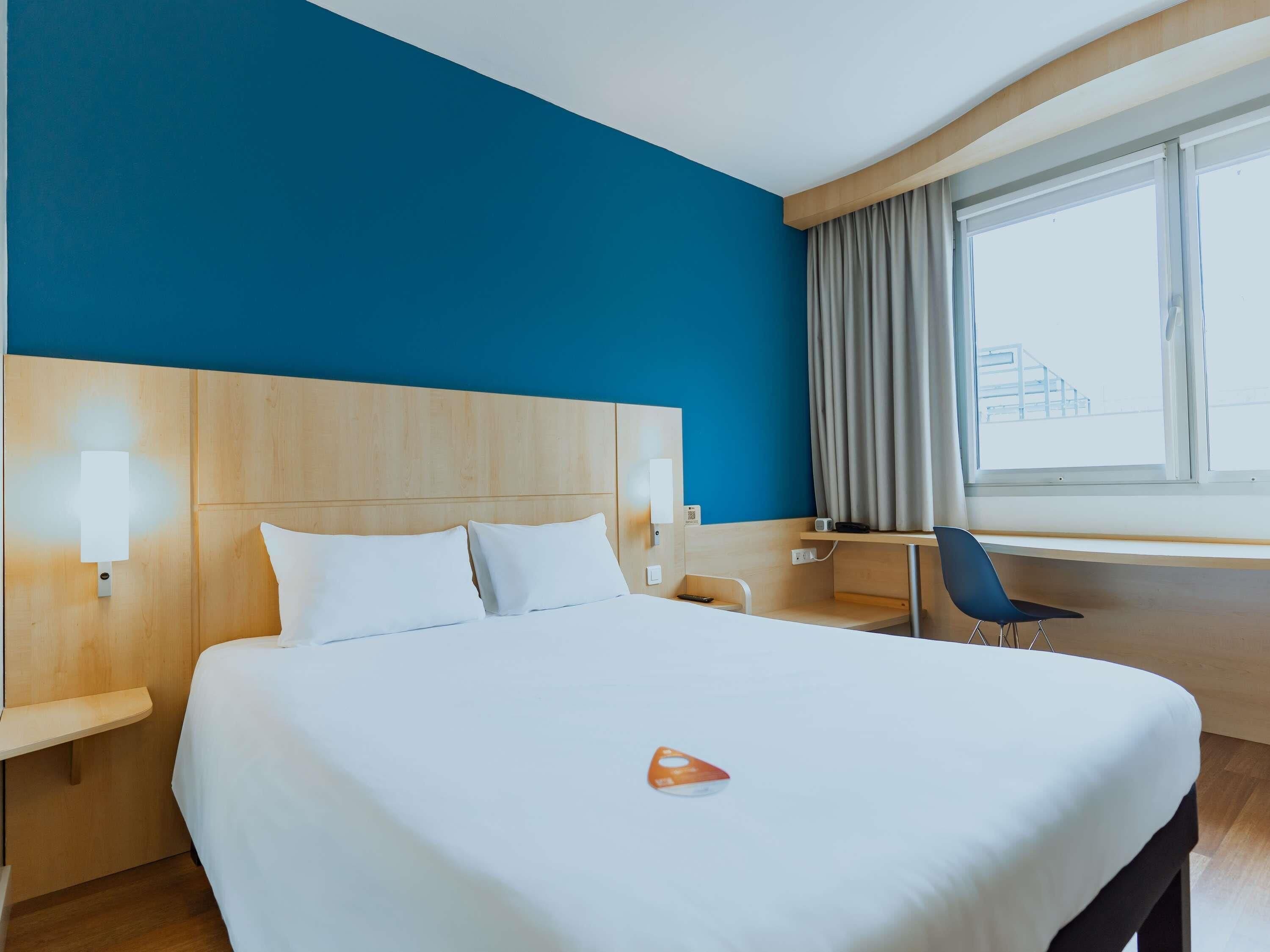 A hotel room with a large bed, a desk, a chair, a window with curtains, and a view of the outside.