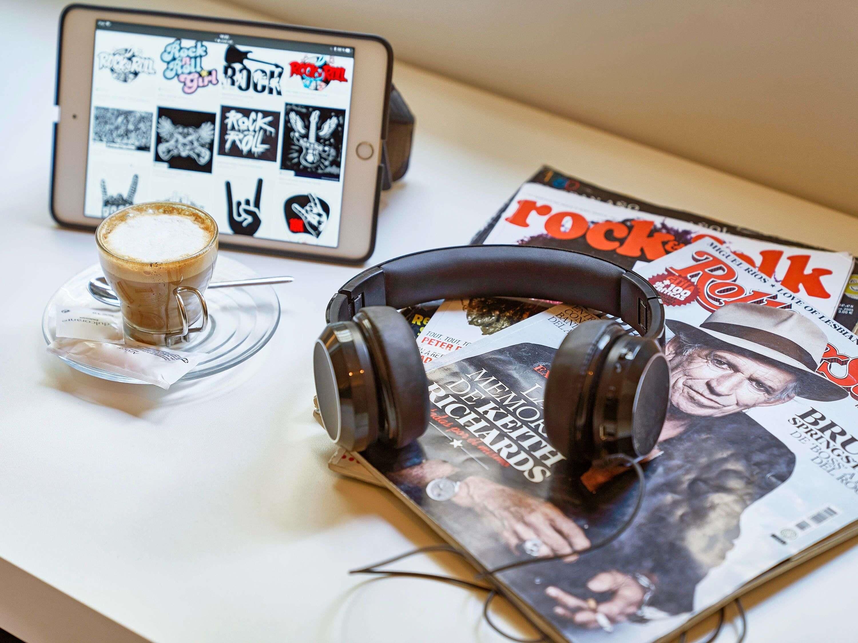 Una tableta mostrando gráficos de música rock, un par de audífonos, una taza de café y una revista sobre una mesa.