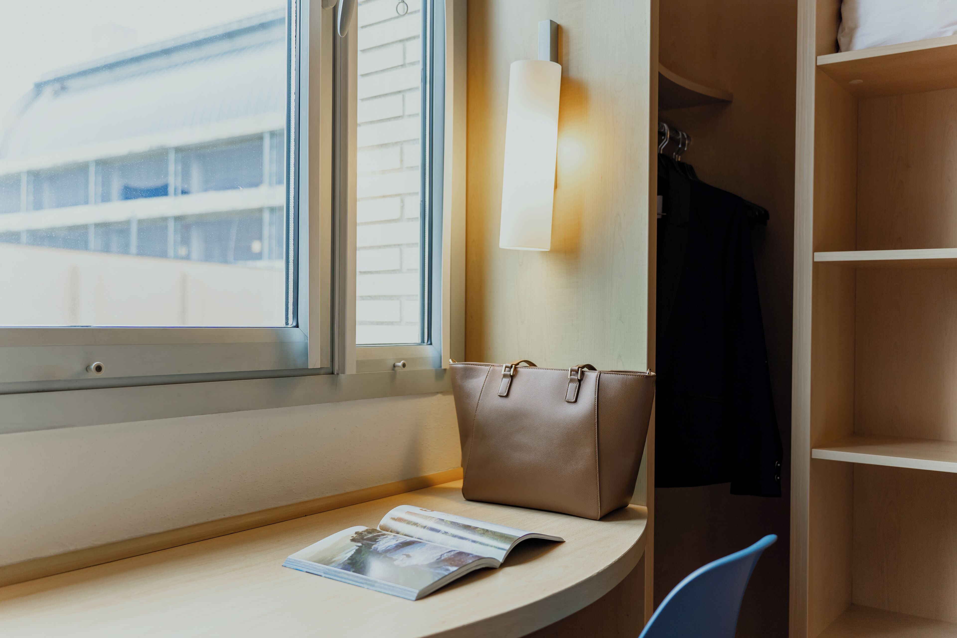 A room with a window, a table with a magazine, a brown handbag, and a coat rack with a black coat.
