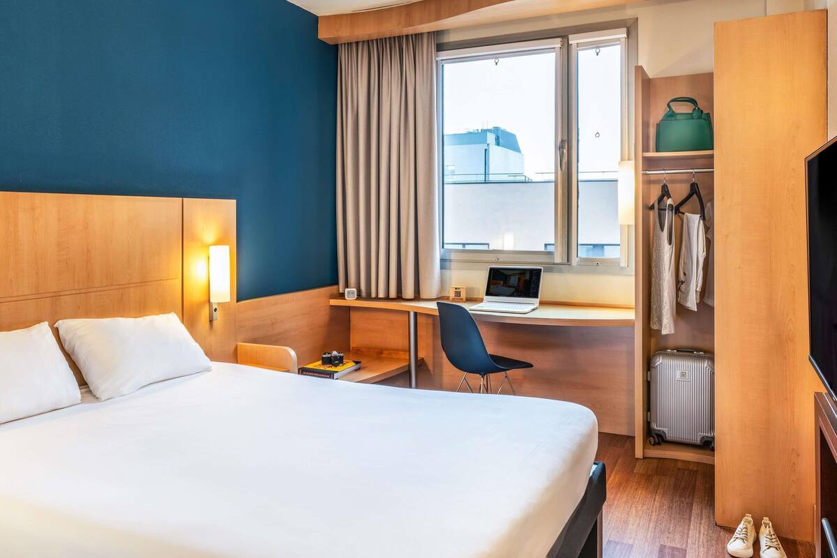 Quarto de hotel com cama, escrivaninha, laptop, cadeira, guarda-roupa e mala.