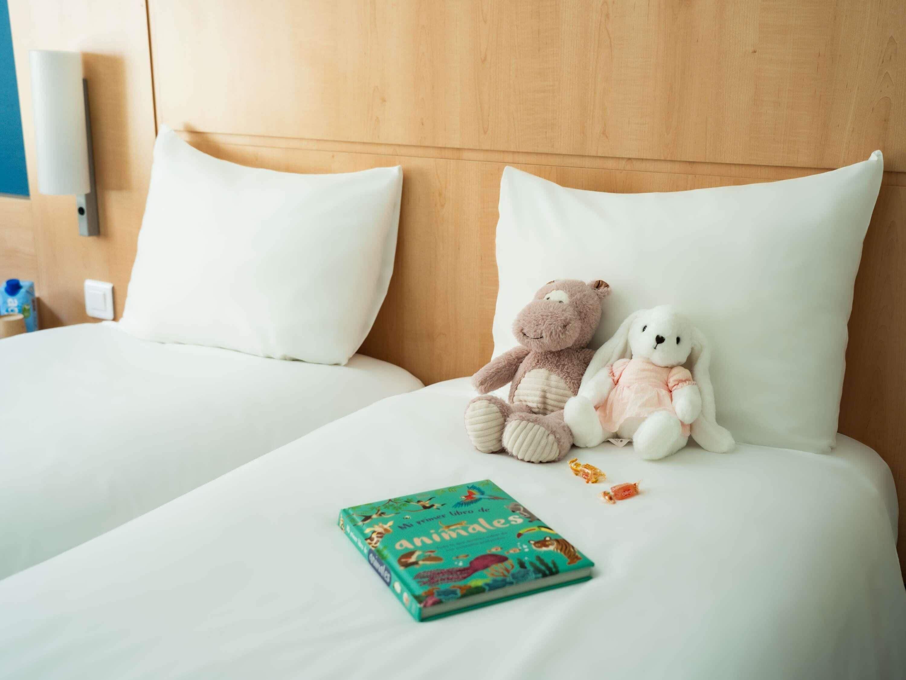 Ein Hotelzimmer mit zwei Betten, weißer Bettwäsche, einem Stofftier, einem Buch und einem kleinen Snack.