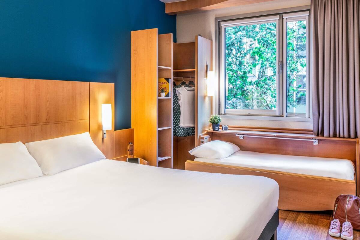 Quarto de hotel com uma cama grande, uma cama menor, um guarda-roupa, uma janela com vista para árvores e uma mesa de cabeceira com abajur.