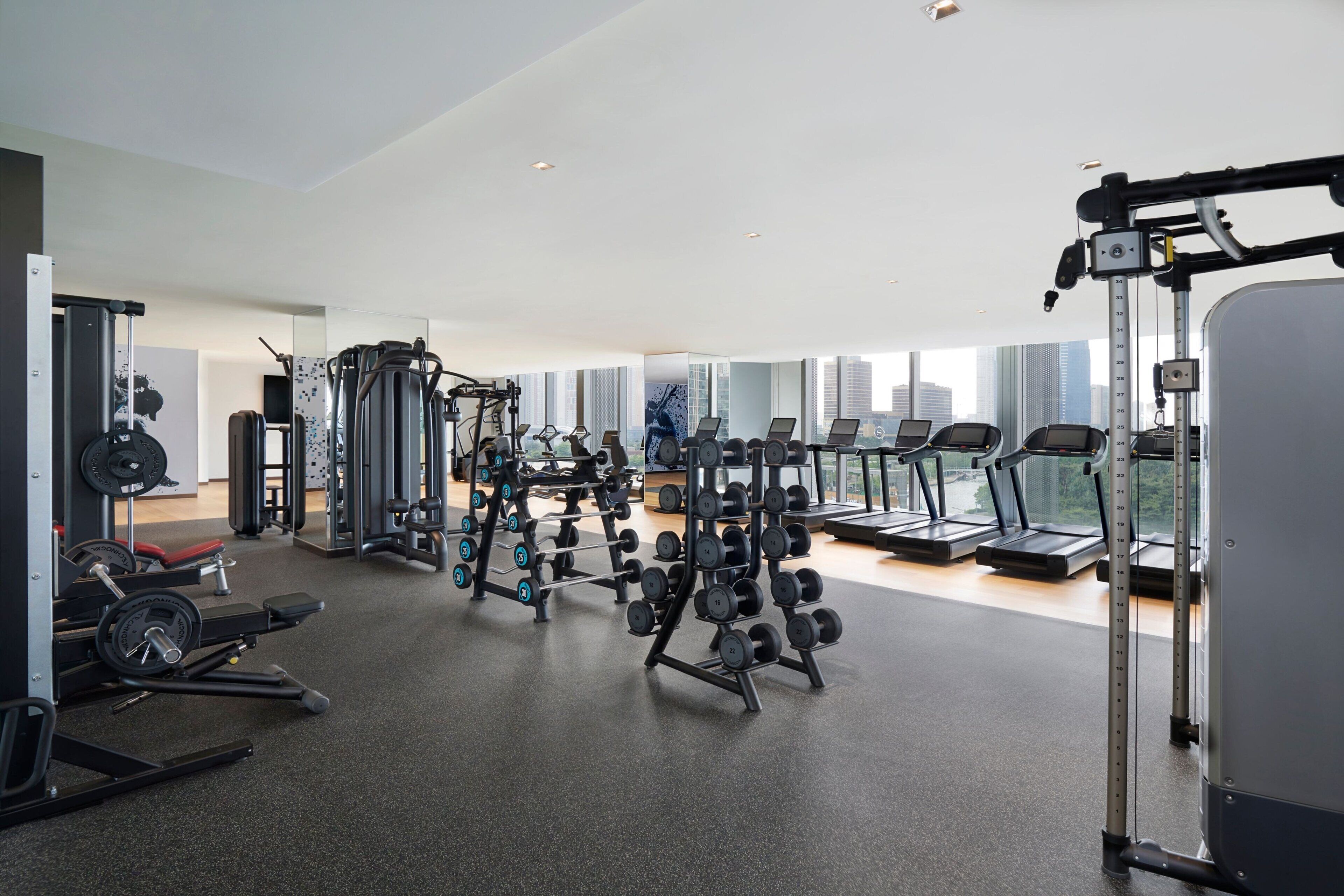 A modern gym with various exercise equipment, large windows, and a city view.