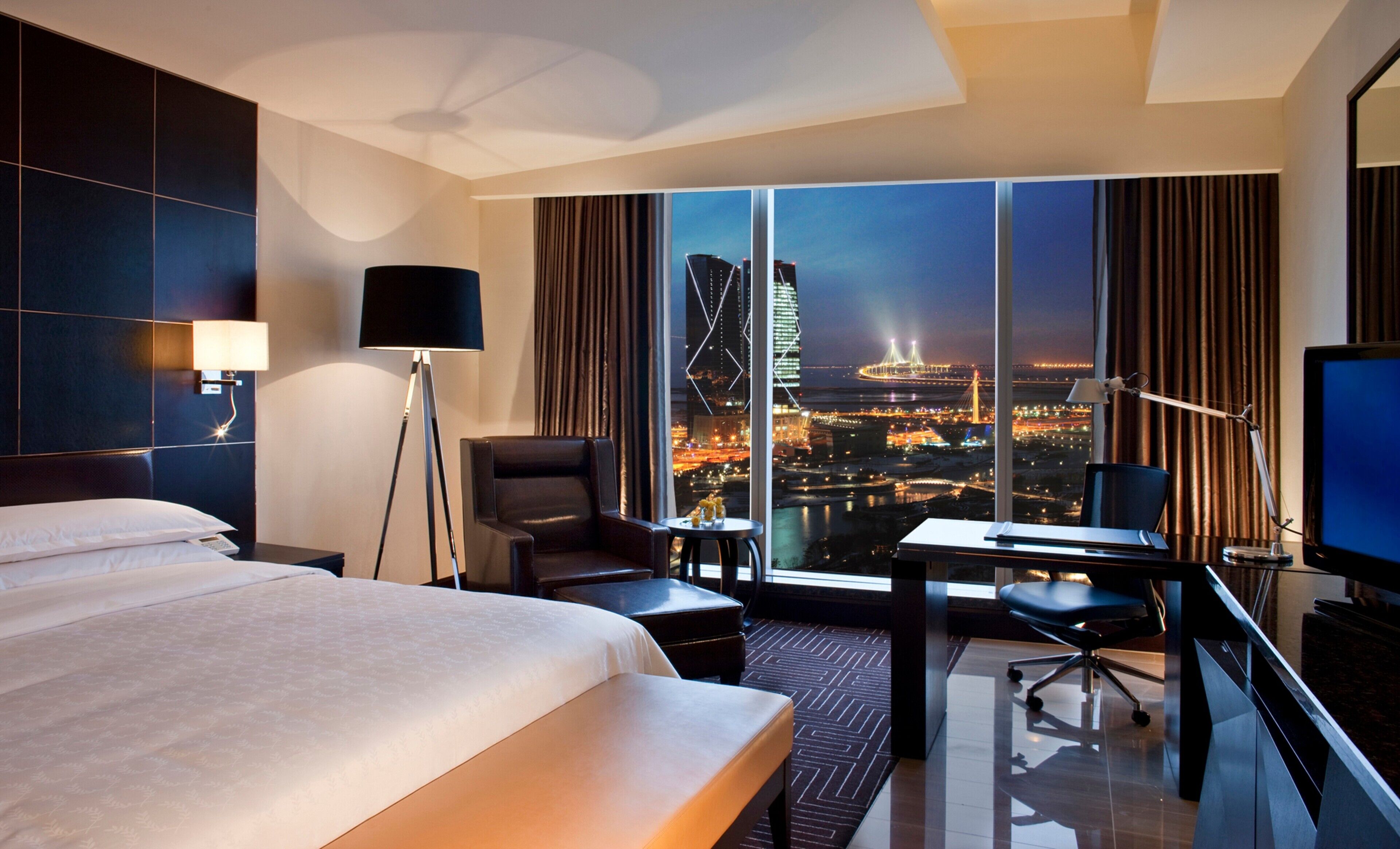Une chambre d’hôtel avec un grand lit, un bureau avec une chaise, une télévision et une vue sur le paysage urbain, avec ses bâtiments illuminés.