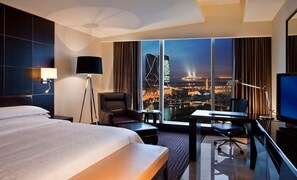 A hotel room with a large bed, a desk with a chair, a TV, and a view of a cityscape with illuminated buildings.