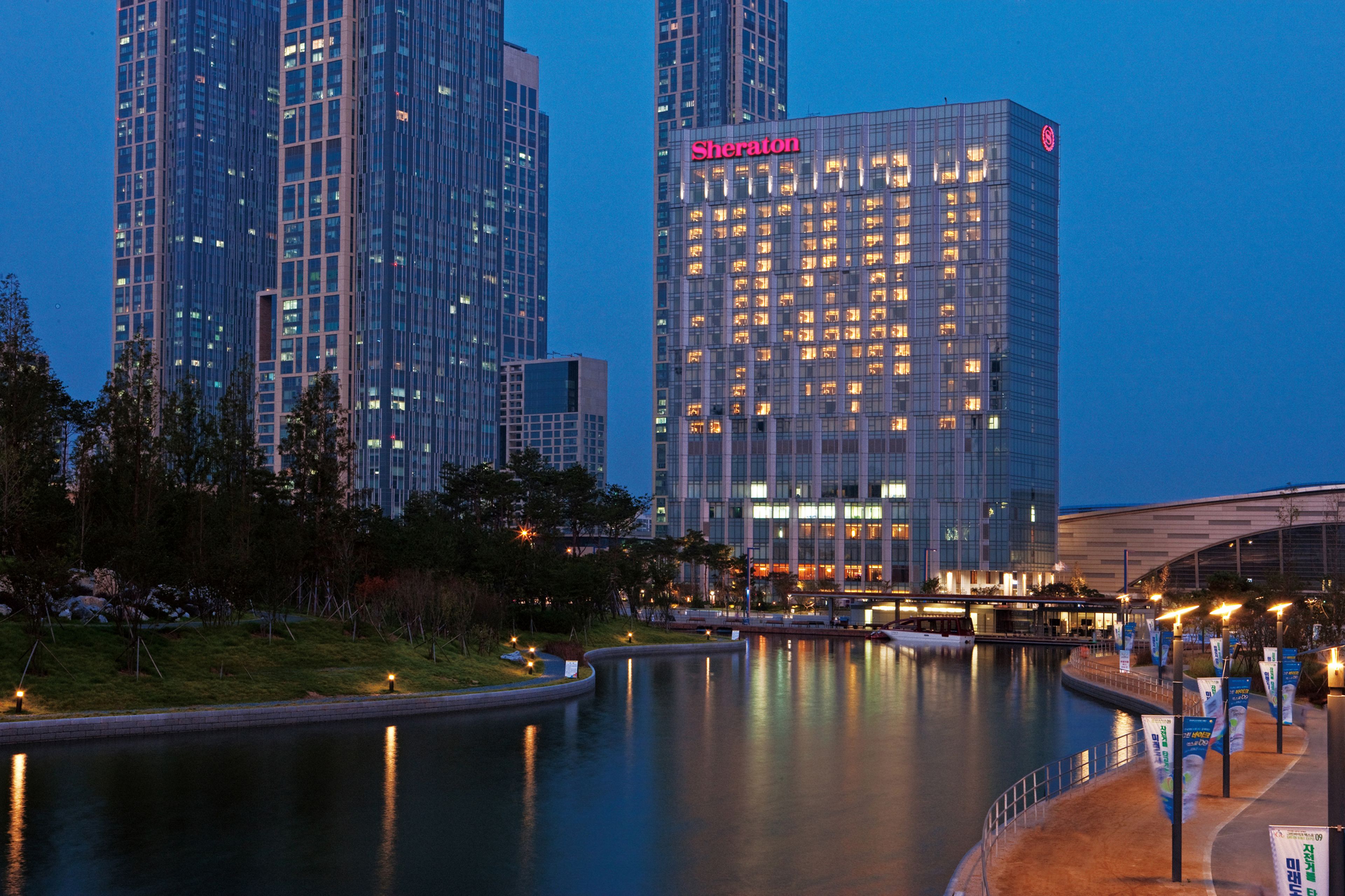 Un hotel Sheraton con fachada de vidrio moderna, reflejado en el agua, y un paisaje urbano circundante.