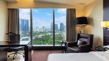 A hotel room with a large window, a desk with a chair, a bed, a side table, a lamp, and a telephone.