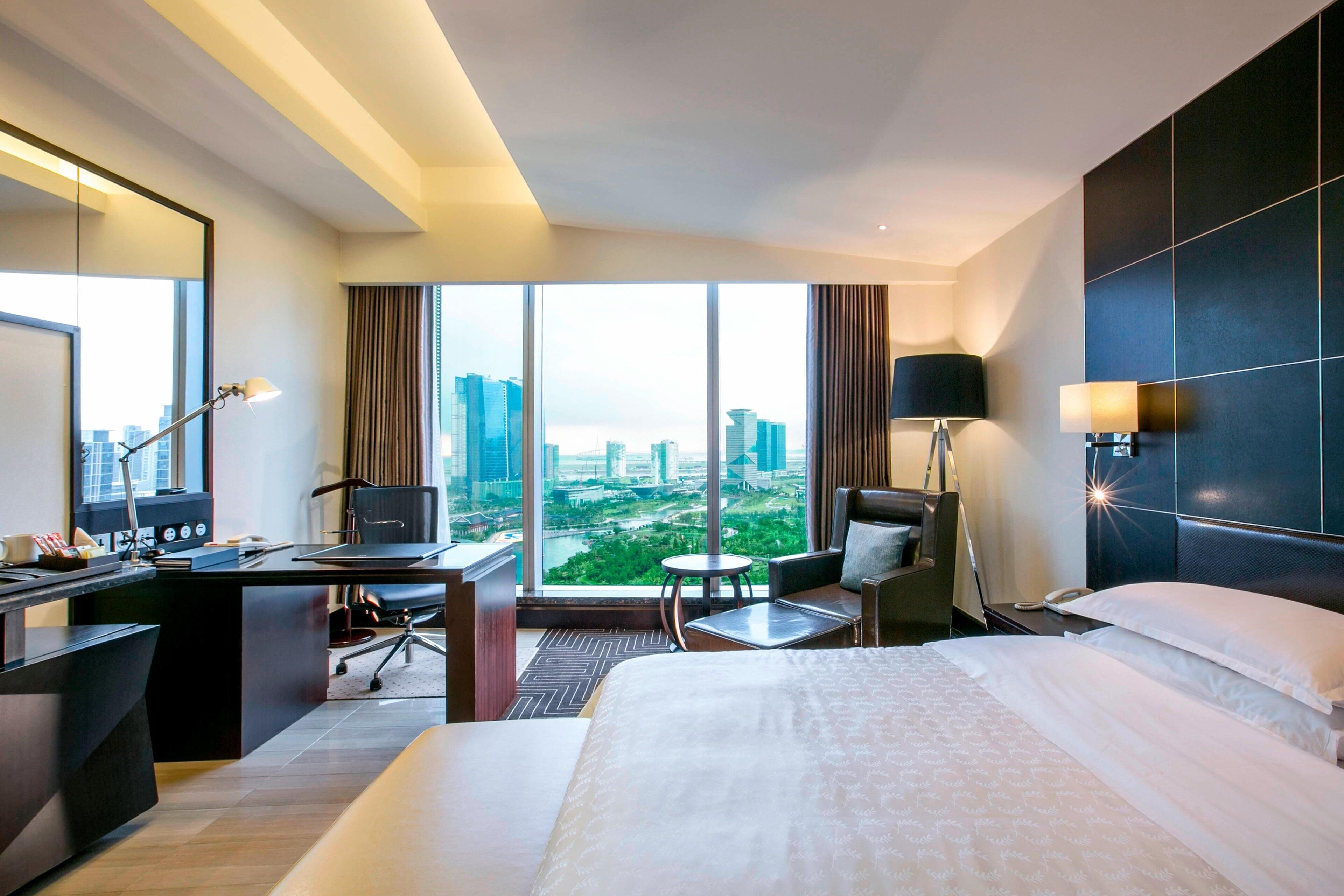 A hotel room with a large bed, a desk, a chair, a small table, and a view of a cityscape through large windows.