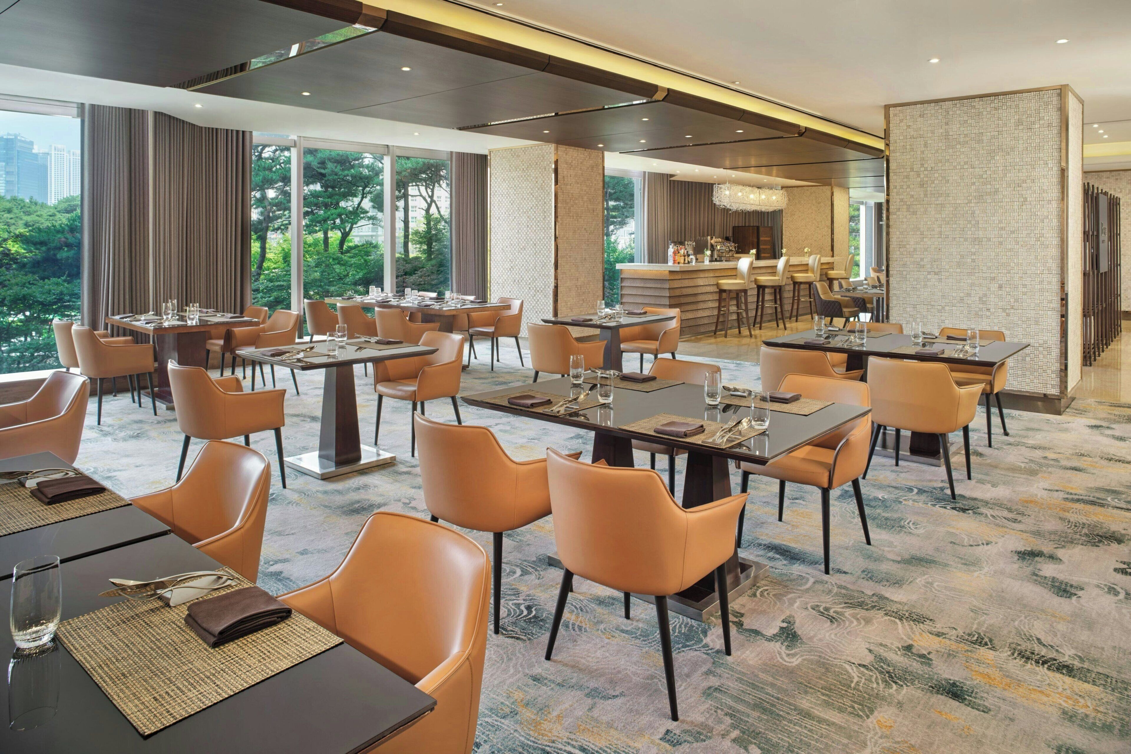 A hotel dining area with multiple tables and chairs, a bar, and large windows offering a view of the outdoors.