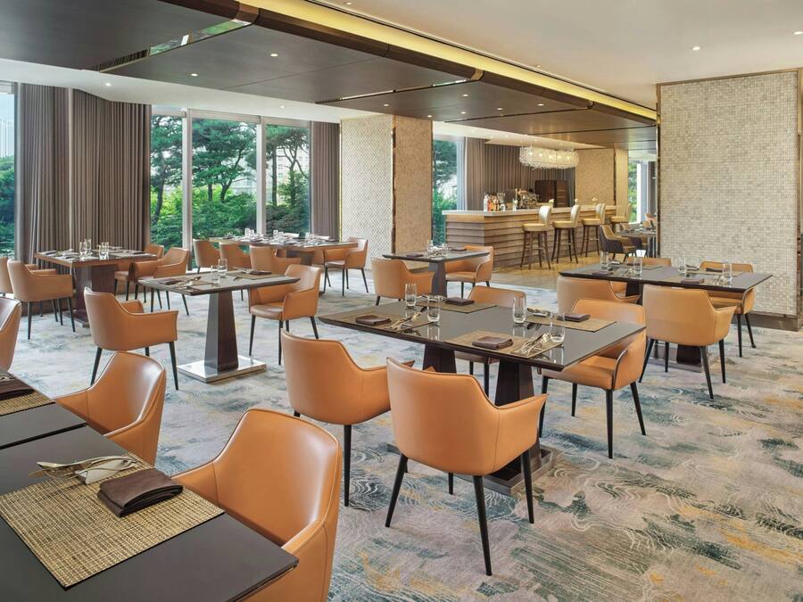 A hotel dining area with multiple tables and chairs, a bar, and large windows offering a view of the outdoors.