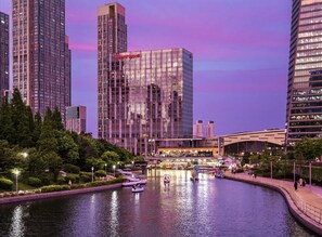 A modern hotel with a glass facade, surrounded by a water canal with boats and a pedestrian walkway.