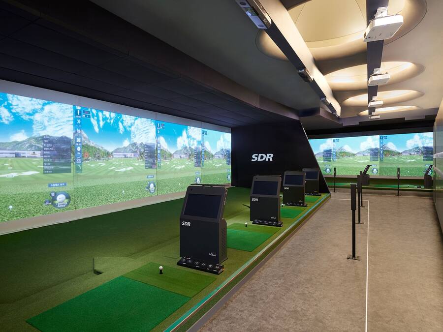 Un área moderna de simulación de golf en interiores con varias pantallas mostrando campos de golf, una zona de césped verde y varios dispositivos electrónicos de seguimiento de puntuación identificados como 'SDR'.