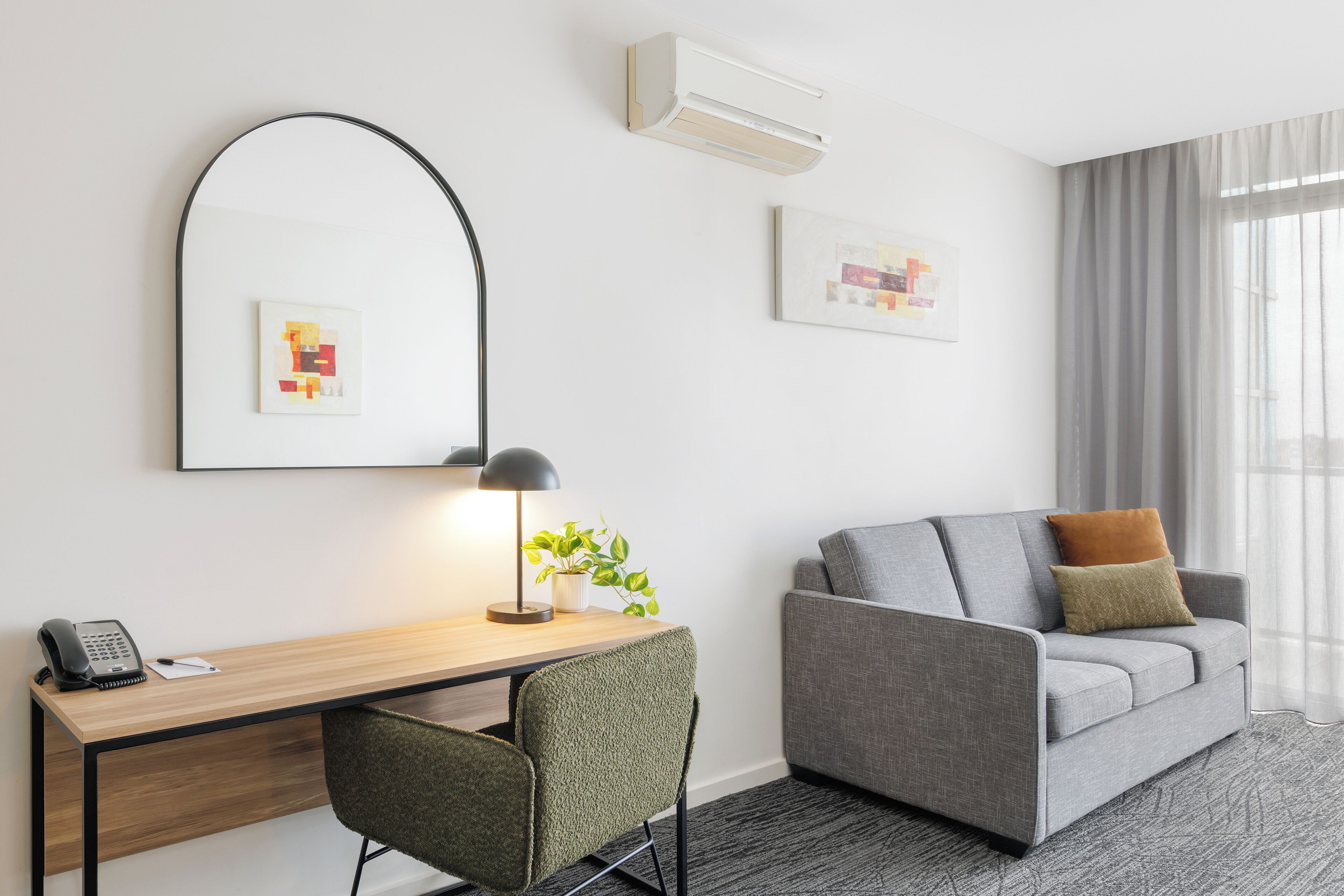 Habitación de hotel moderna con un escritorio de madera, una silla verde, un sofá gris con dos cojines, un espejo grande y dos cuadros enmarcados en la pared.