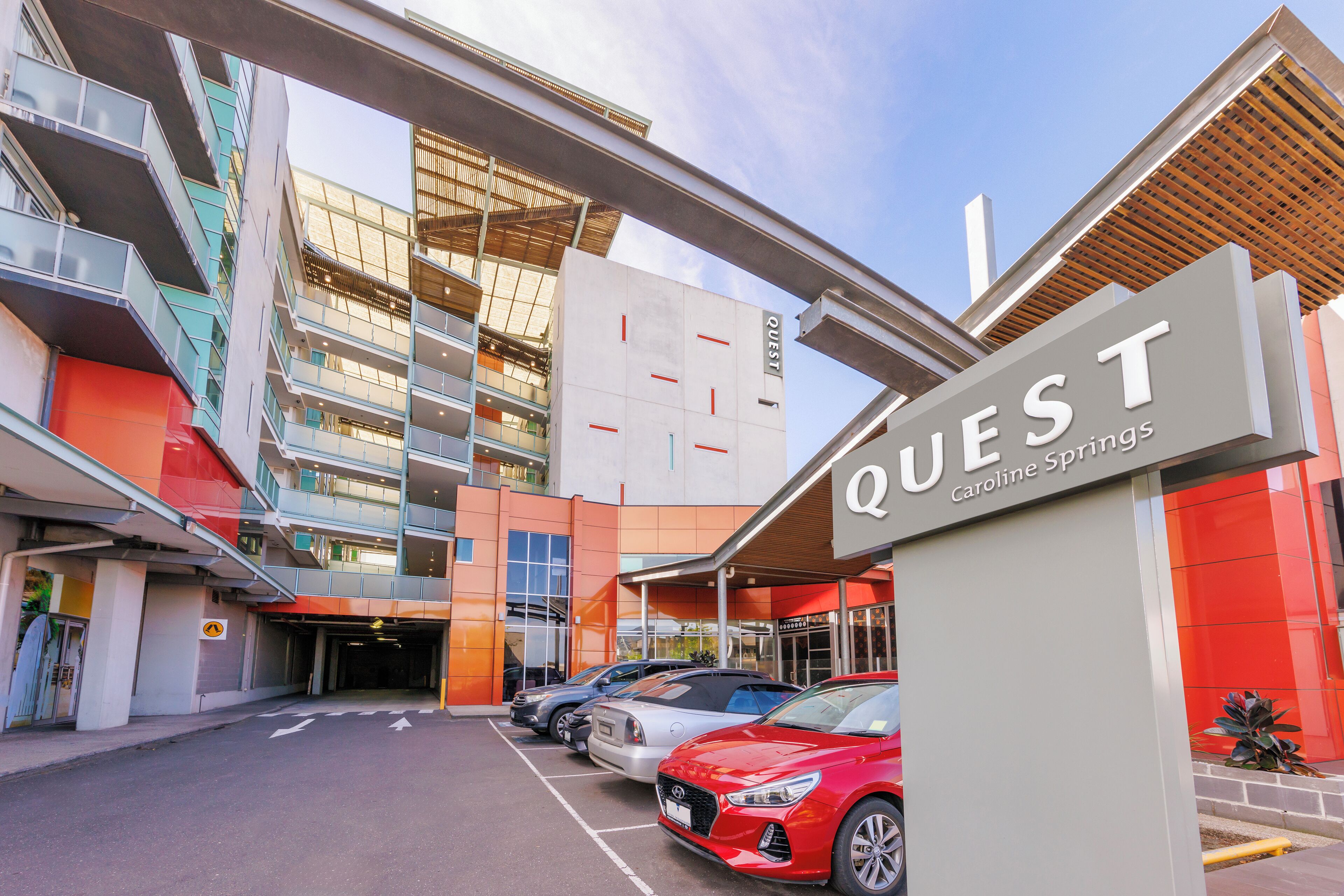 A modern hotel entrance with a sign reading "QUEST Caroline Springs." There are multiple cars parked in front, and the building features a multi-story structure with balconies.