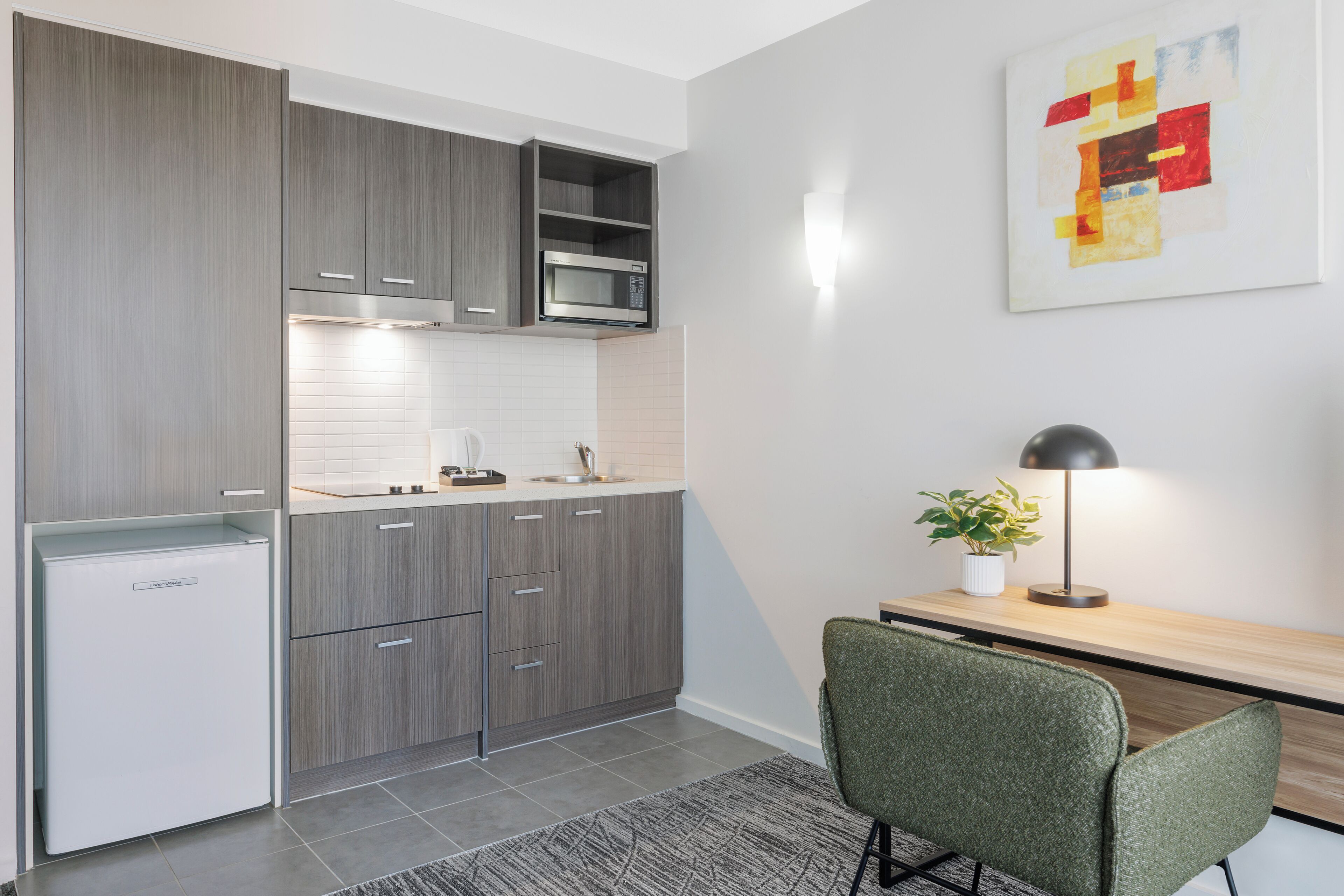 A modern kitchen with grey cabinets, a white refrigerator, and a microwave. There's a dining area with a green chair and a table, and a lamp on the table.