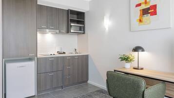 A modern kitchen with grey cabinets, a white refrigerator, and a microwave. There's a dining area with a green chair and a table, and a lamp on the table.