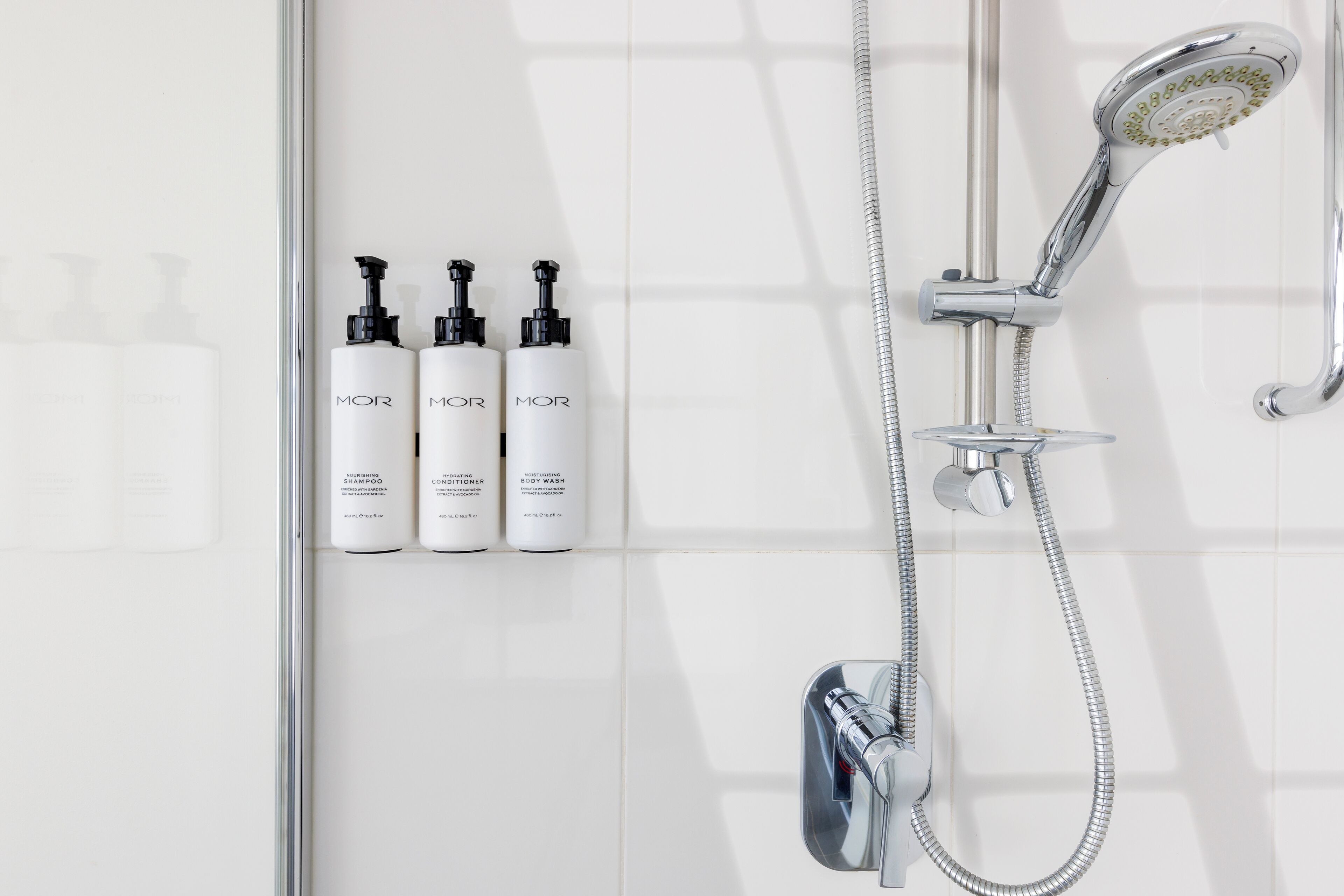 A modern bathroom with a chrome showerhead, a handheld showerhead, and three bottles of MOR body wash and conditioner.