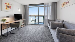 A modern hotel room with a grey sofa, a desk with a flat-screen TV, a small table with a chair, and a balcony with a view of the city. - Quest Caroline Springs (Caroline Springs)