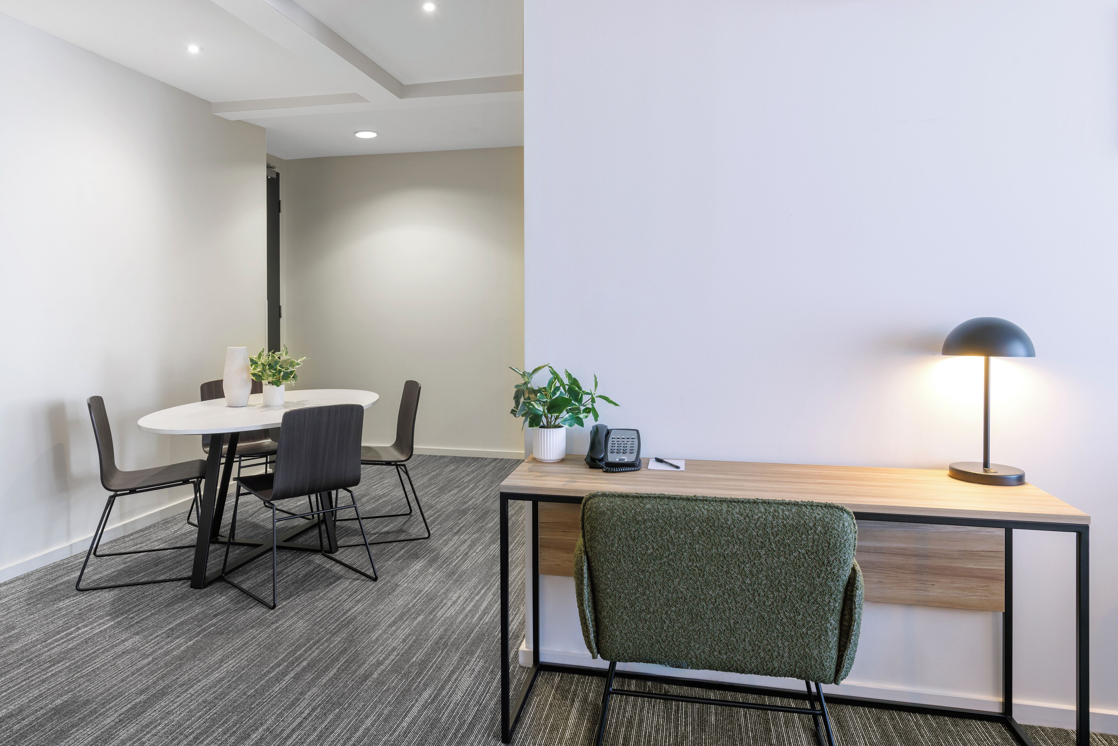 A modern office space with a round table, chairs, a desk with a computer, a lamp, and a potted plant.