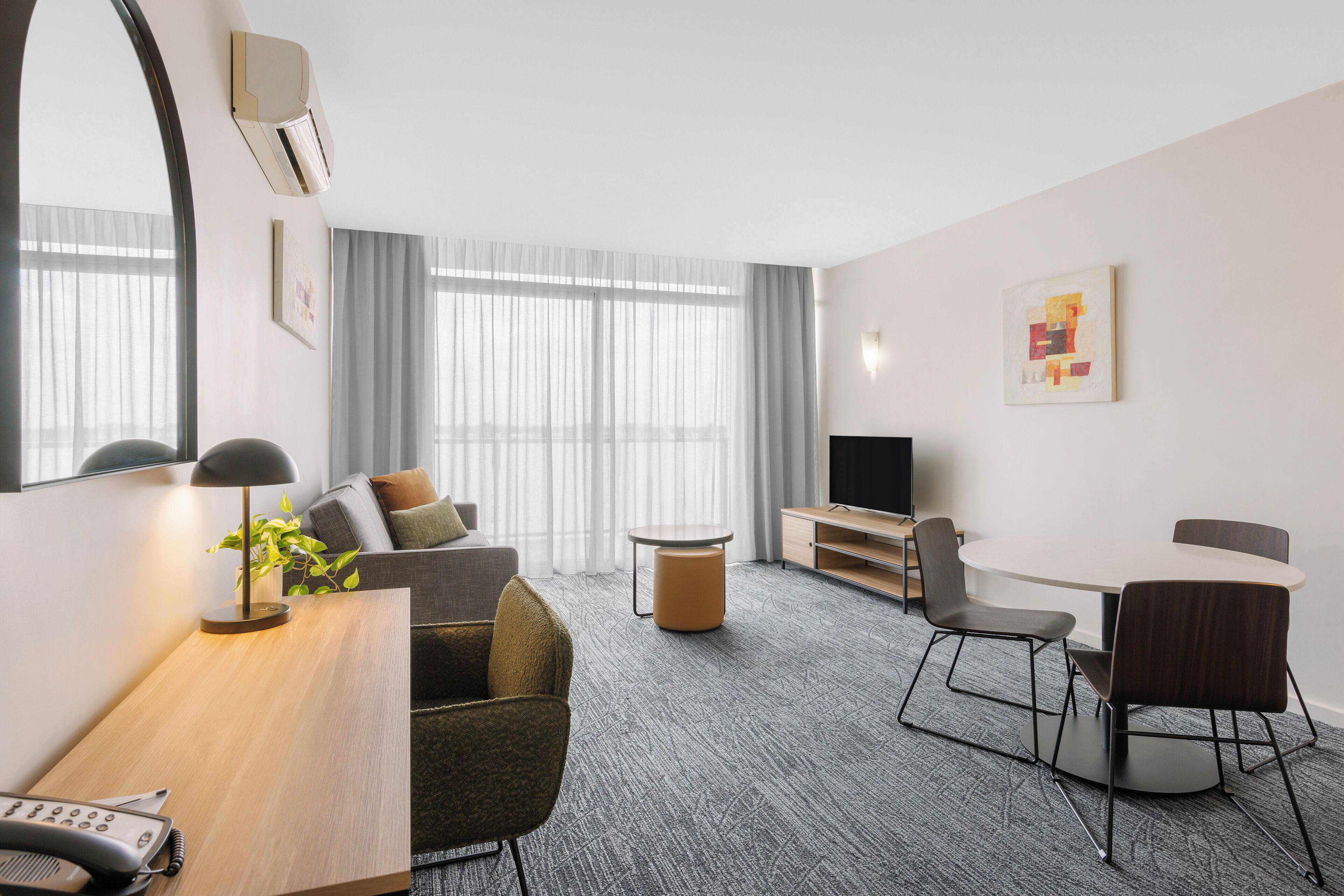 A modern hotel room with a sofa, a dining table with chairs, a TV, and a large window with curtains.