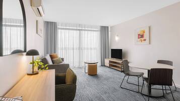 A modern hotel room with a sofa, a dining table with chairs, a TV, and a large window with curtains.