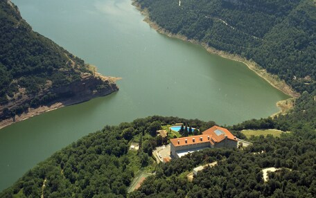 A large body of water surrounded by forested hills with a building featuring a red roof and a swimming pool.