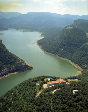 A large body of water surrounded by forested hills with a building featuring a red roof and a swimming pool.