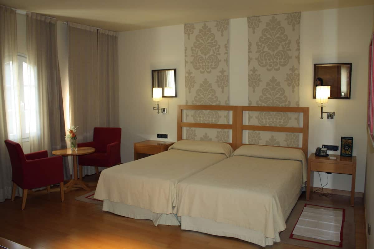 Quarto de hotel com duas camas, uma escrivaninha, uma cadeira e uma pequena mesa com um vaso de flores.