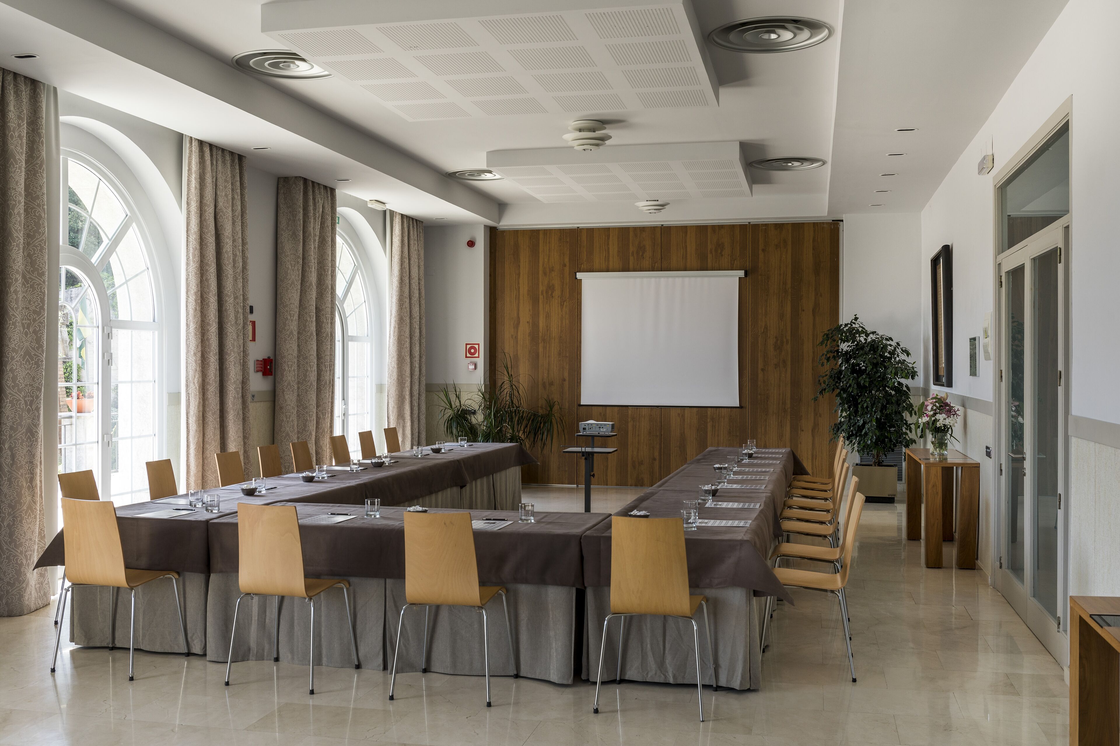 Un salón de conferencias con una larga mesa, sillas, una pantalla de proyector y grandes ventanas con cortinas.