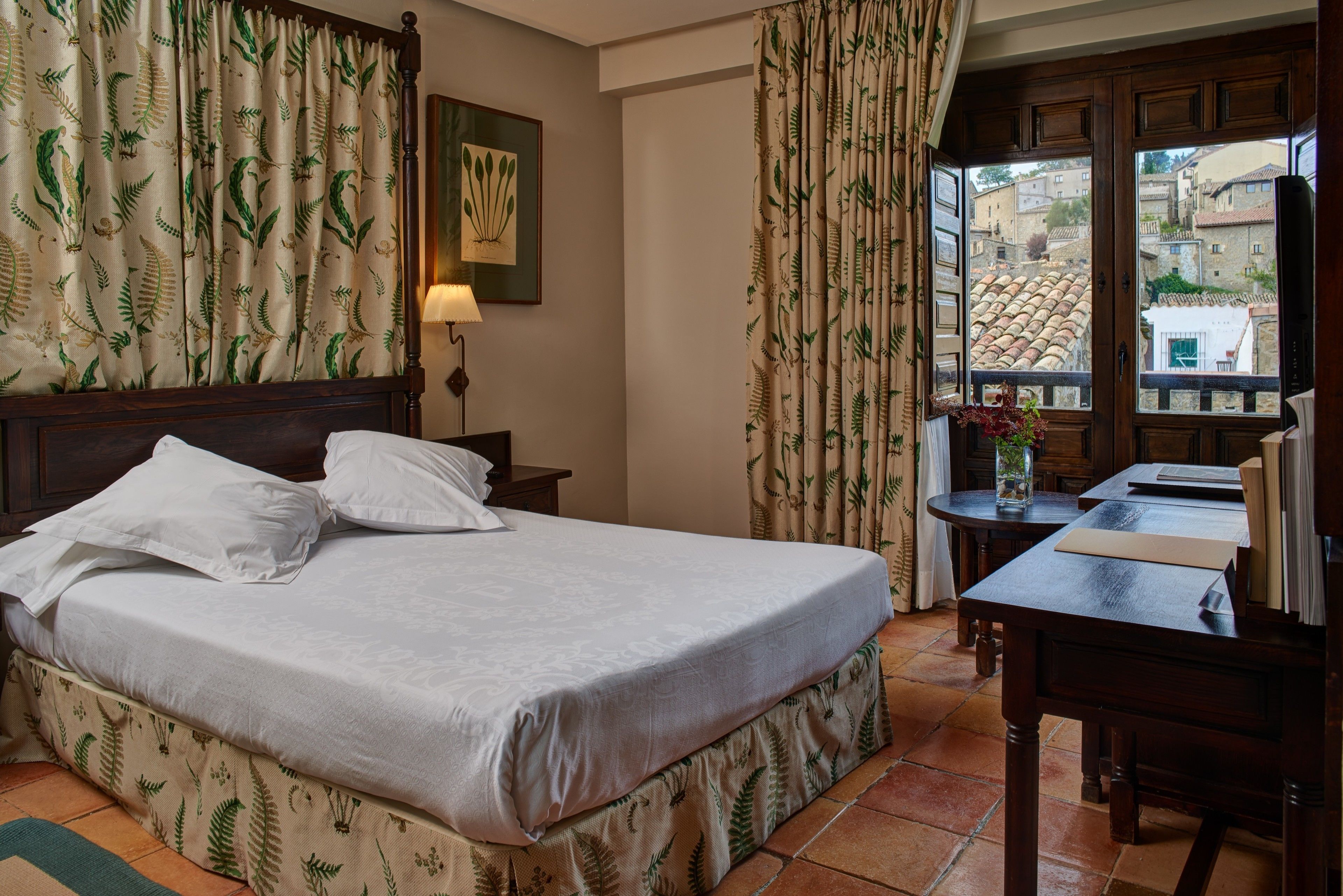 Habitación de hotel con cama, escritorio, lámpara y vistas al pueblo a través del ventanal.