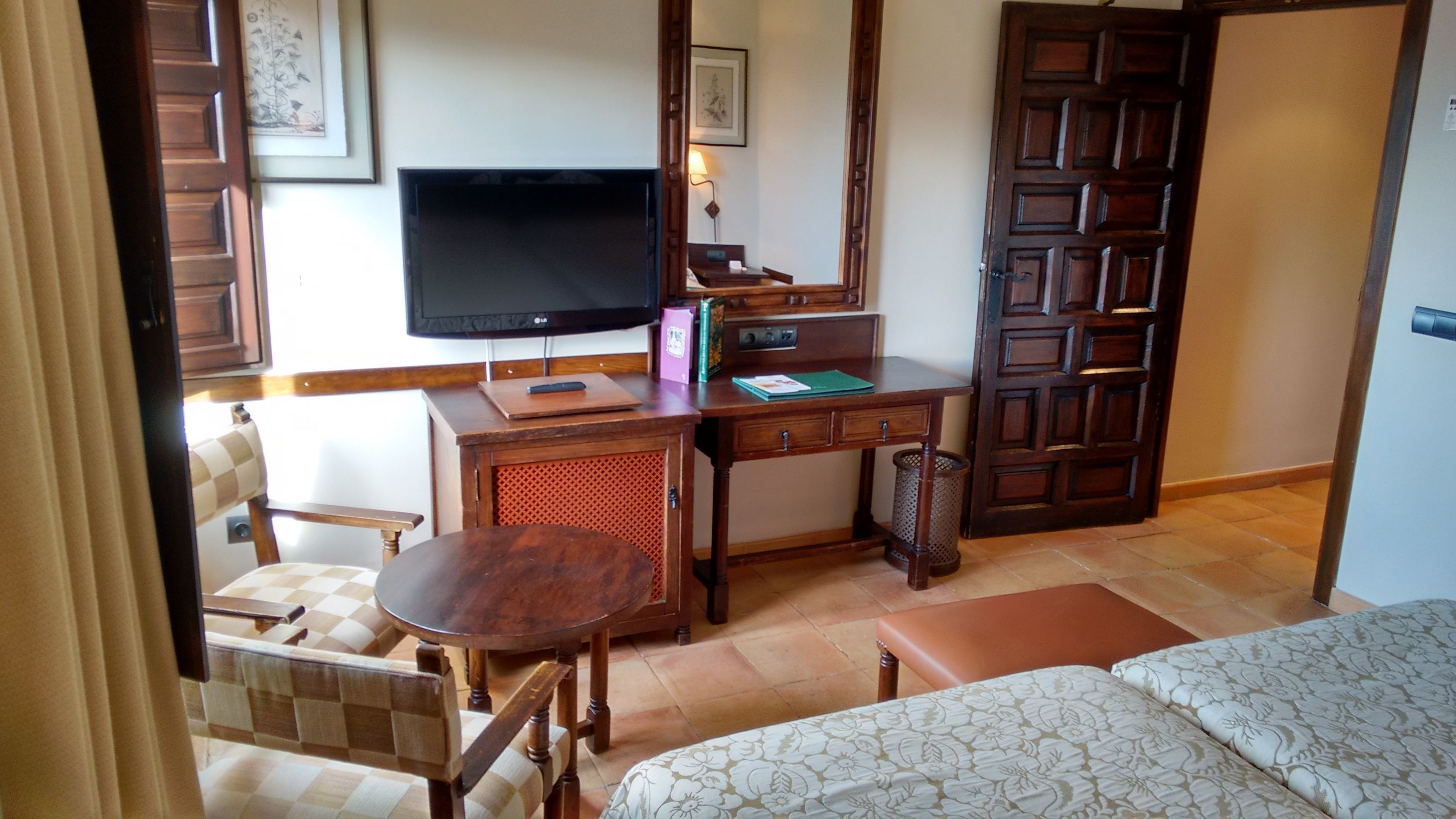 A hotel room with a bed, a desk with a television, a chair, a small table, and a door.