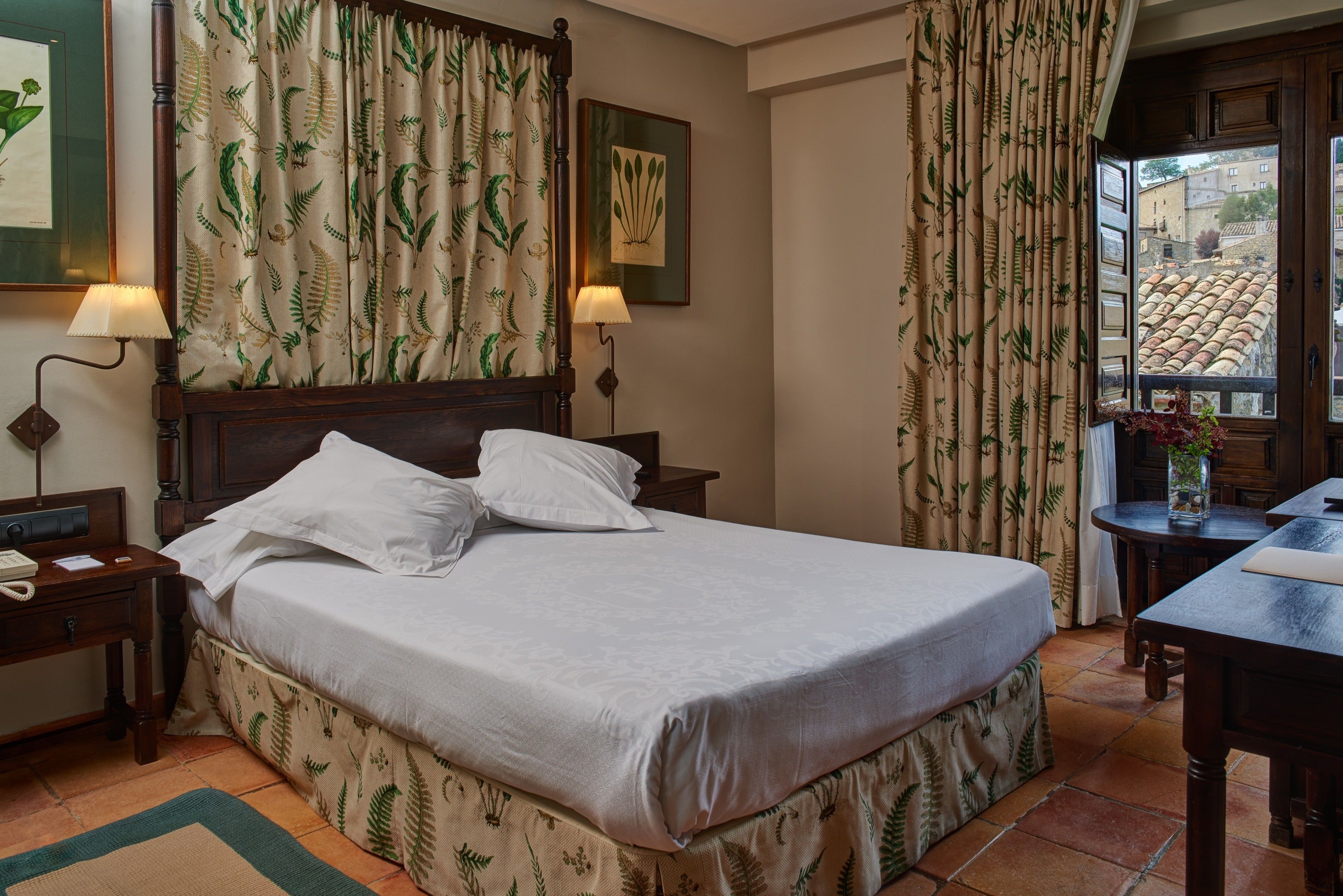 A neatly made bed with a white bedspread and matching pillows, flanked by two bedside lamps. A wooden nightstand with a telephone is to the left. The room features a large window with a view of buildings and a tiled roof outside.