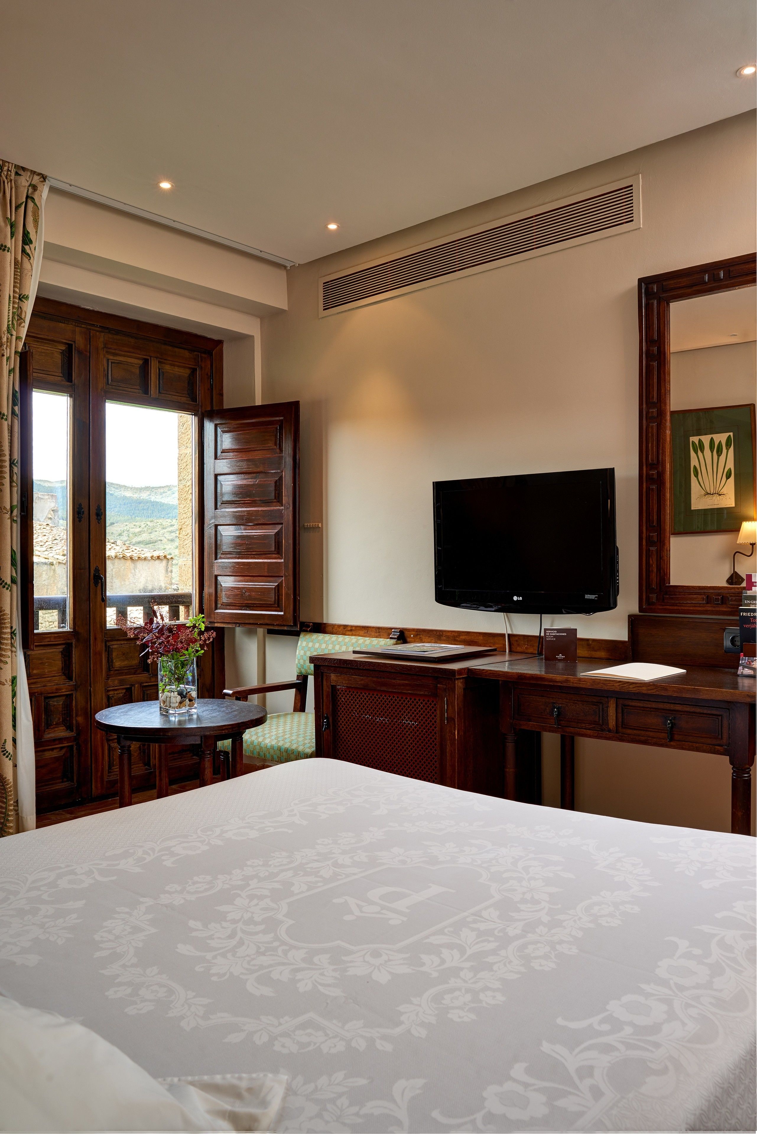 A hotel room with a large bed, a desk with a flat-screen TV, a chair, a small table with a vase, and a window with curtains.