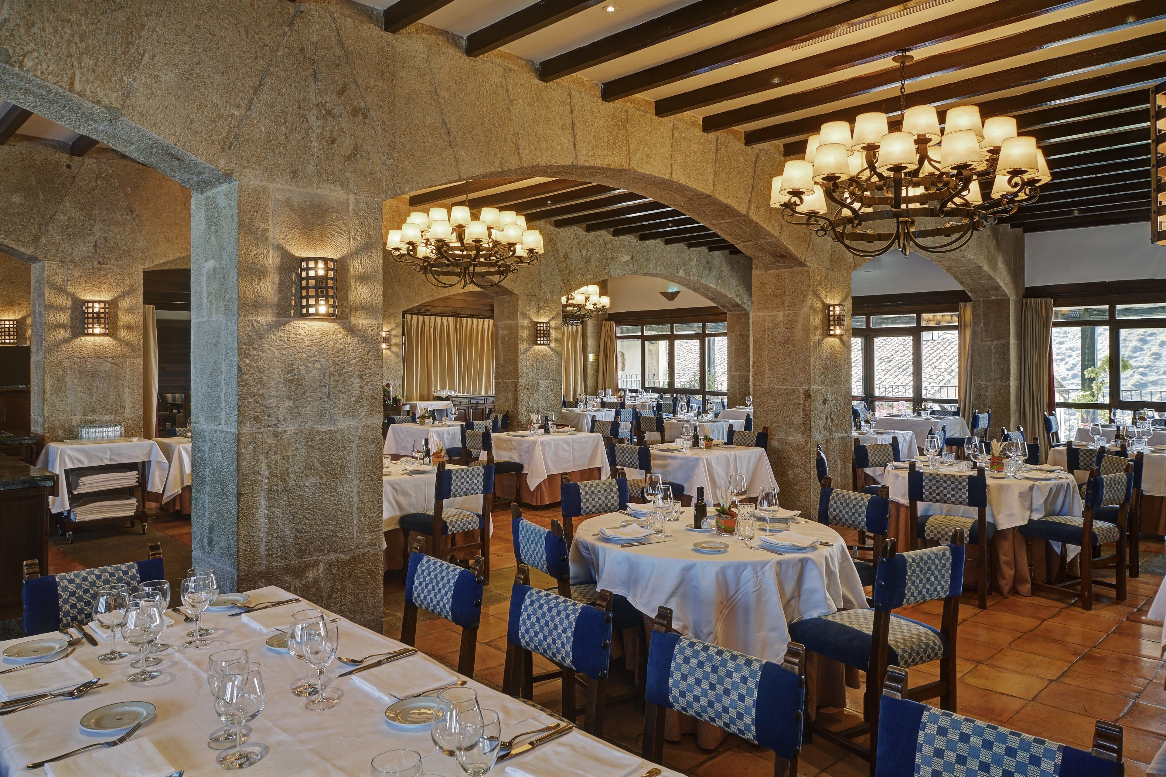 Un restaurante con varias mesas dispuestas para comer, con respaldos de sillas a cuadros y manteles blancos.