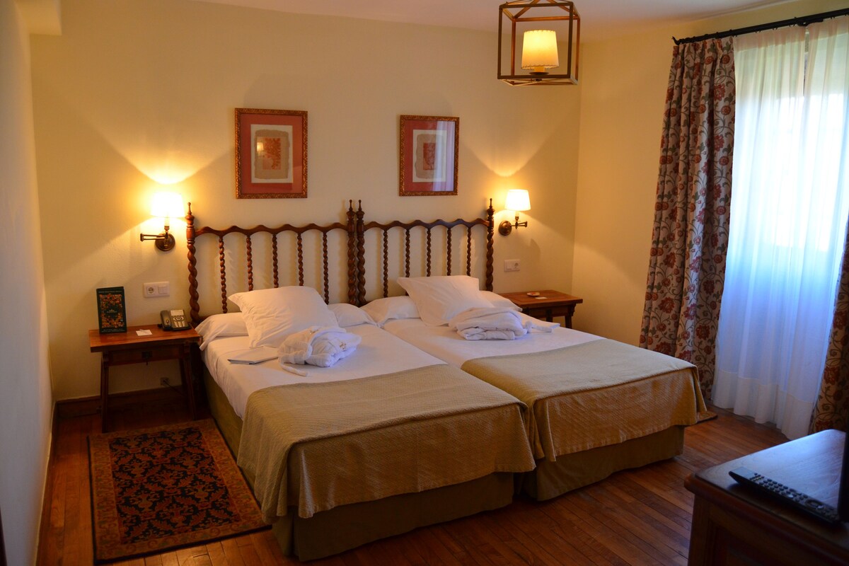 A hotel room with two beds, a wooden headboard, bedside lamps, a nightstand, a small table, a remote control, and a floral curtain.