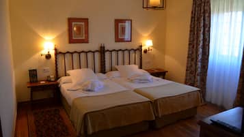 A hotel room with two beds, a wooden headboard, bedside lamps, a nightstand, a small table, a chair, and a window with curtains.