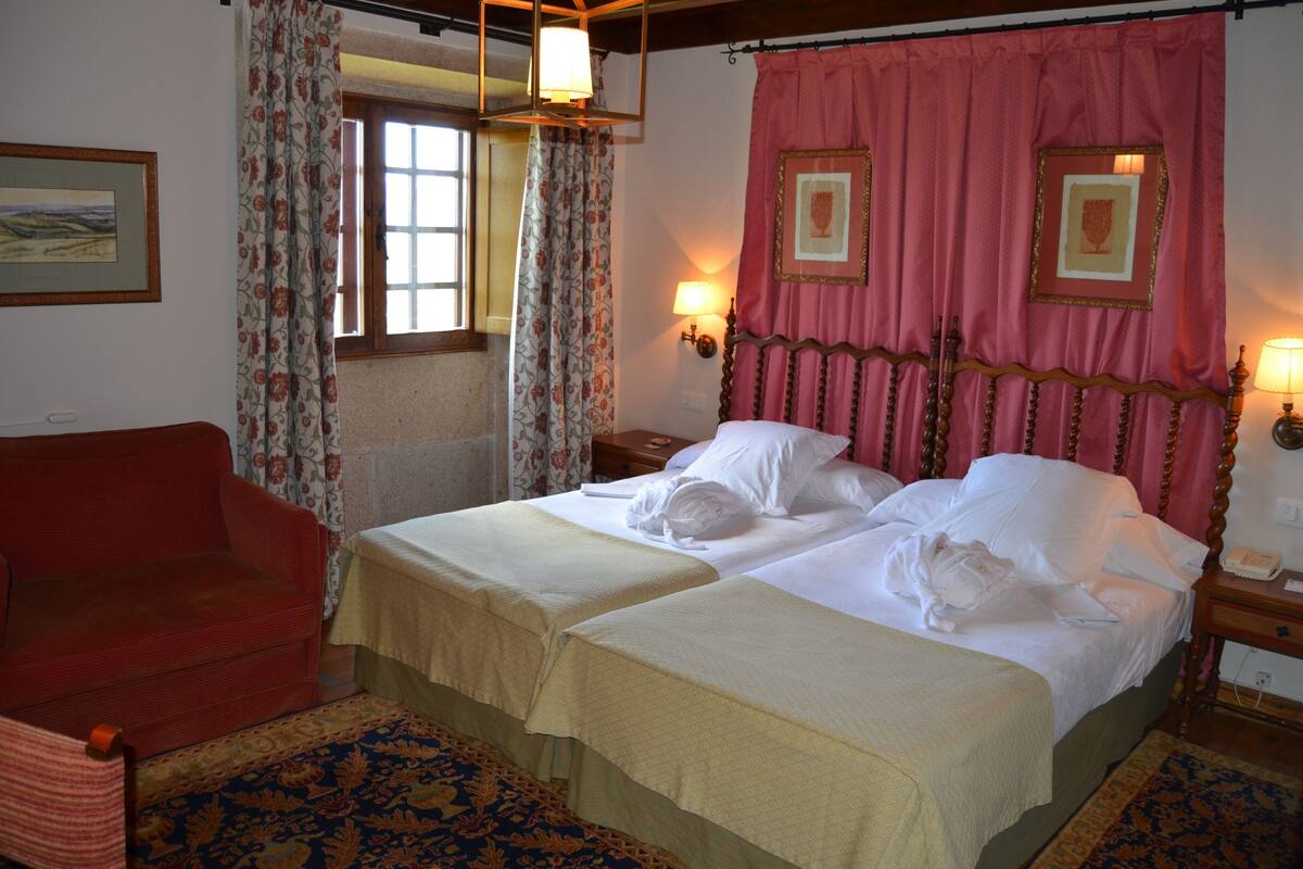 A hotel room with two beds, a red armchair, a patterned rug, and a window with floral curtains.