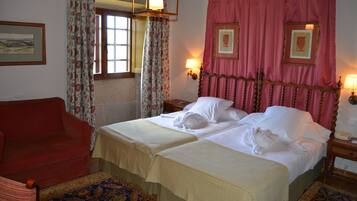 Habitación de hotel con dos camas, un sillón rojo, un tapete estampado y una ventana con cortinas florales.