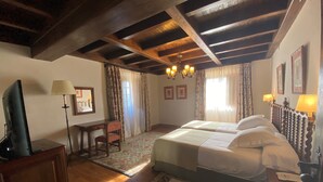 A hotel room with a wooden ceiling, a bed with white linens, a desk with a lamp, and a television.